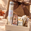 A curated gift box set styled in an autumn setting. The set includes a small wooden box containing a clear bottle of bath salts, three decorative soap bars, a knitted scarf, and a wrapped item, all presented in a burlap tote bag with a sheer brown ribbon.