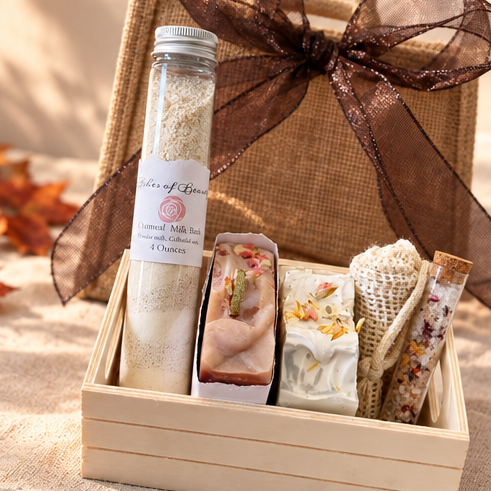 A curated gift box set styled in an autumn setting. The set includes a small wooden box containing a clear bottle of bath salts, three decorative soap bars, a knitted scarf, and a wrapped item, all presented in a burlap tote bag with a sheer brown ribbon.